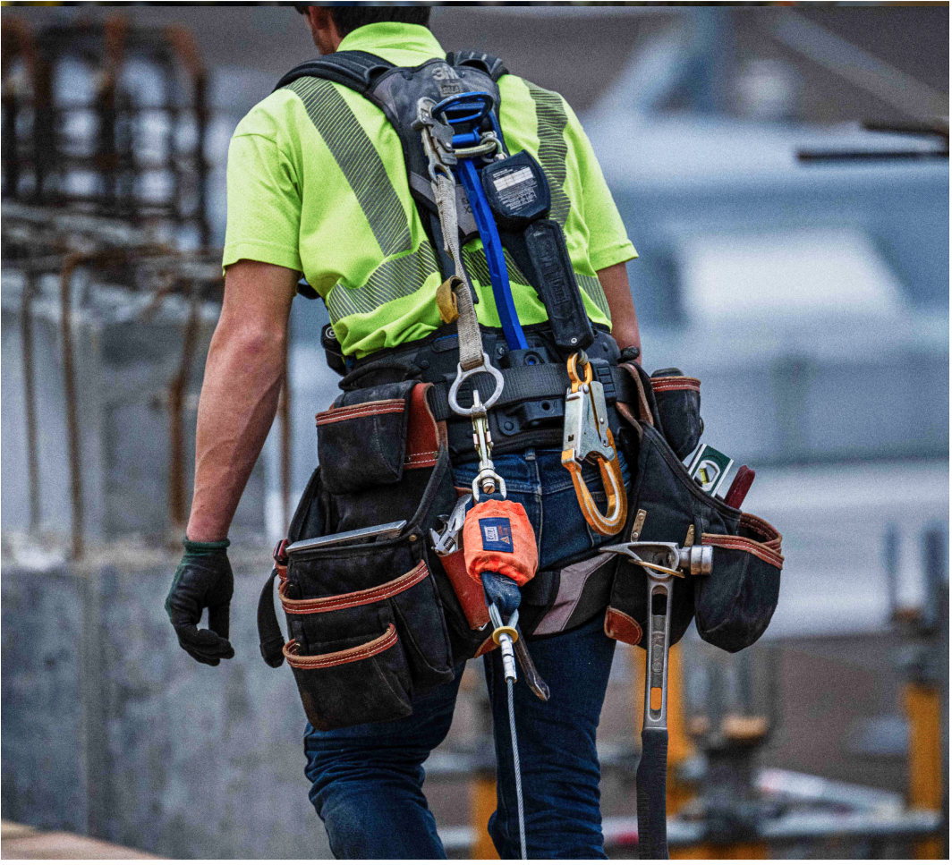 Image of a construction worker from behind wearing a yellow high visibility shirt, fall protection harness and other safety gear.