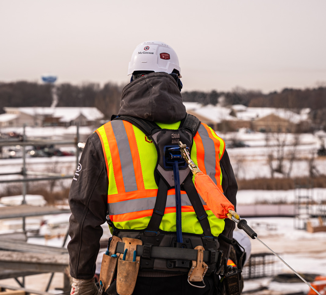 Image of a construction worker from behind wearing a yellow high visibility shirt, fall protection harness and other safety gear.